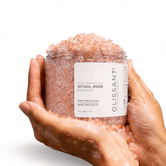 A clear plastic jar filled with pink Himalayan bath salts, held in two hands against a clean white background. The label reads 'GLISSANT ROSE + GERMANIUM OIL RITUAL ROSÉ BATH SALTS'.