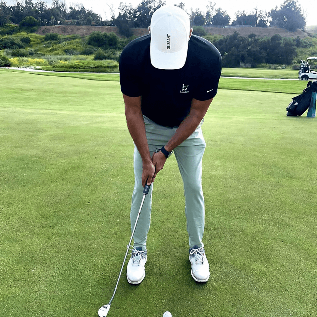 A man on a golf course wearing a black Nike polo shirt, light green pants, white Nike golf shoes, and a white cap, preparing to hit a golf ball with a putter.