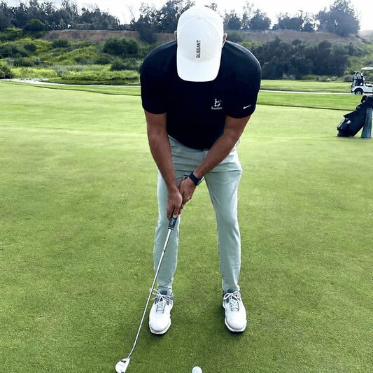 A man on a golf course wearing a black Nike polo shirt, light green pants, white Nike golf shoes, and a white cap, preparing to hit a golf ball with a putter.