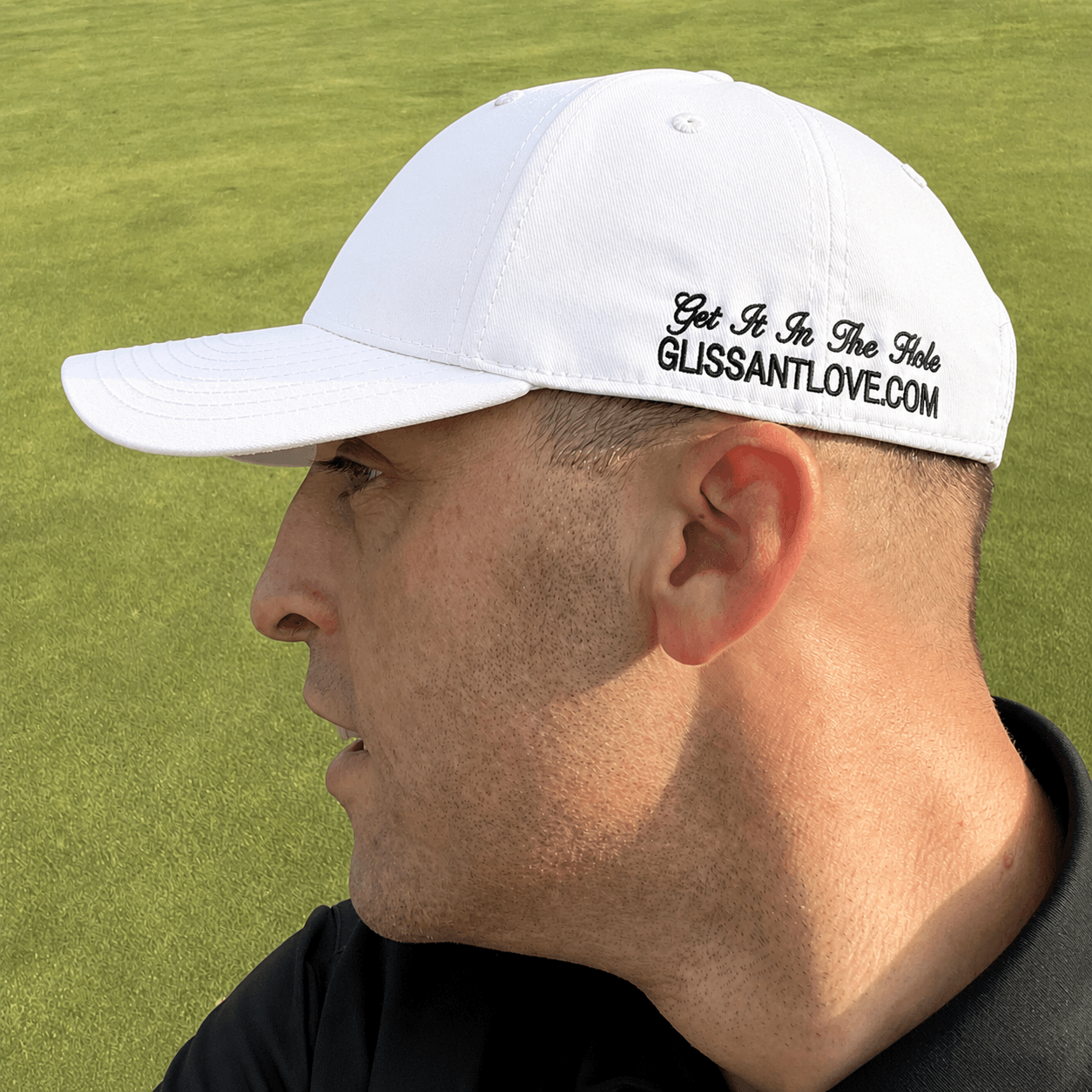 A man in profile wearing a white baseball cap with black embroidered text reading 'Get It In The Hole GLISSANTLOVE.COM' on a green grass background.